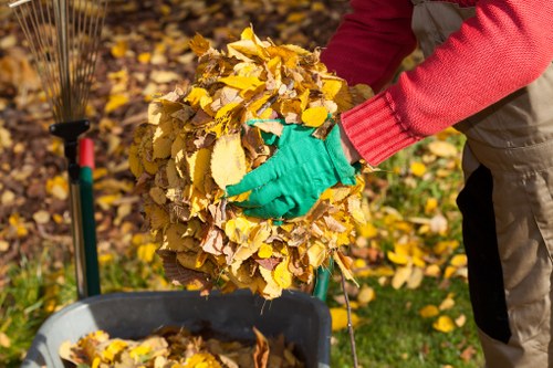 A person wearing red long-sleeved clothing and bright teal gardening gloves is shown collecting a large bundle of yellow and brown fallen autumn leaves. The individual is holding the bundle with both hands over a grey wheelbarrow, which is partially visible in the foreground. The background includes a garden area with more fallen leaves scattered on the grass and a garden rake with a red and green handle leaning against a shrub or plant. The scene appears to be outdoors during daytime, likely in a suburban garden or yard in the UK, with natural light illuminating the leaves and the person's attire. This image relates to rubbish removal services provided by Office Clearance Beckenham, exemplifying garden clearance activities typical of waste management tasks in the Beckenham area.