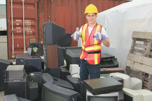 A young male worker wearing a yellow safety helmet, high-visibility vest with red and yellow stripes, and purple gloves, stands outdoors amidst a large pile of discarded electronic appliances and office equipment. The debris includes various types of monitors, desktop computers, keyboards, and other computer accessories, with some items stacked and overlapping on a paved surface. In the background, there is a rusty shipping container and a wooden pallet leaning against a white temporary barrier, suggesting a site for waste clearance or disposal. The worker is smiling and giving a thumbs-up, indicating a successful rubbish collection or clearance operation undertaken by Office Clearance Beckenham in the local area. The scene is well-lit with natural daylight, emphasizing the mix of textures and colours among the electronic waste, with shades of black, grey, white, and brown predominantly visible, set within an environment typical of a commercial or industrial waste management context.