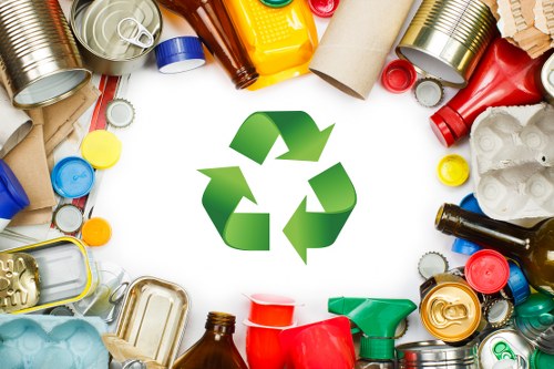 An overhead view of various types of waste materials scattered on a white surface, including metal cans, plastic bottles, glass jars, cardboard tubes, and miscellaneous packaging, surrounding a central green recycling symbol composed of three arrows forming a triangle. The waste items display a range of colours such as silver, green, red, yellow, blue, and brown, with some crushed or flattened and others uncompressed. The image suggests waste segregation and recycling practices relevant to rubbish removal services in Beckenham or the surrounding areas. The arrangement highlights common household or office waste, emphasizing the importance of proper disposal and recycling managed by companies like Office Clearance Beckenham, based in the local postcode area.