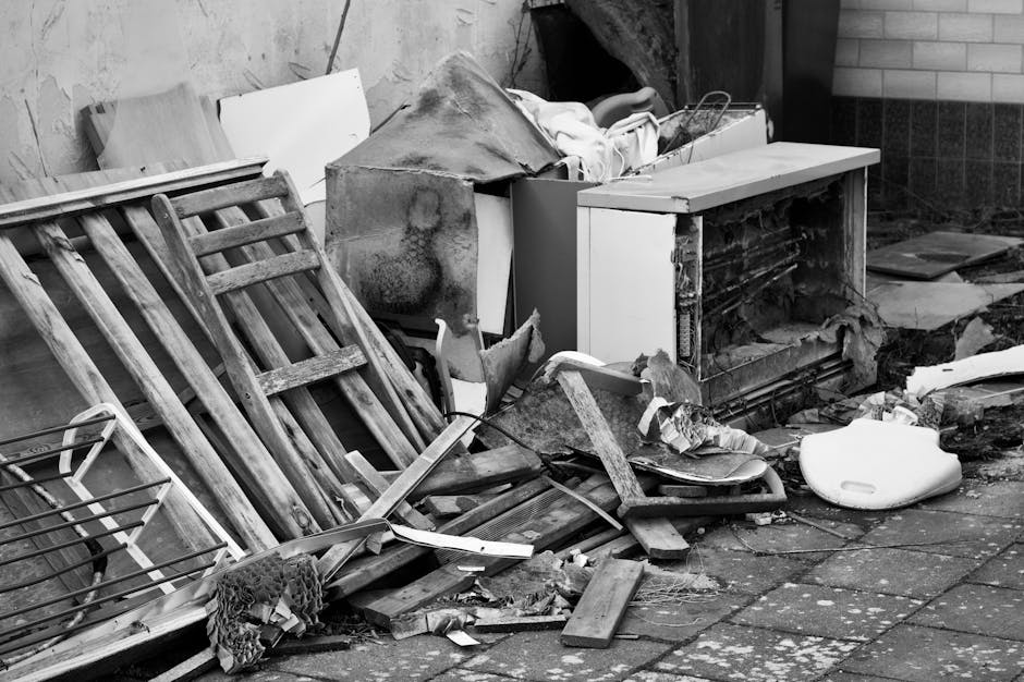 The image displays an outdoor scene with a pile of discarded waste materials placed on a paved surface. At the forefront, several wooden slats and a small metal wire basket are stacked, showing signs
