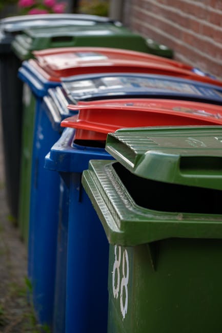 A row of large wheelie bins positioned along a sidewalk beside a brick wall, with the green bin closest to the camera and the others in red, blue, and dark green, extending into the background. The bi